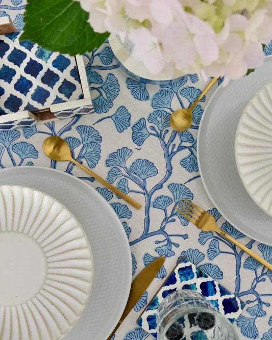 Table setting with blue and white patterned tablecloth, gray plates, gold cutlery, and white flowers.