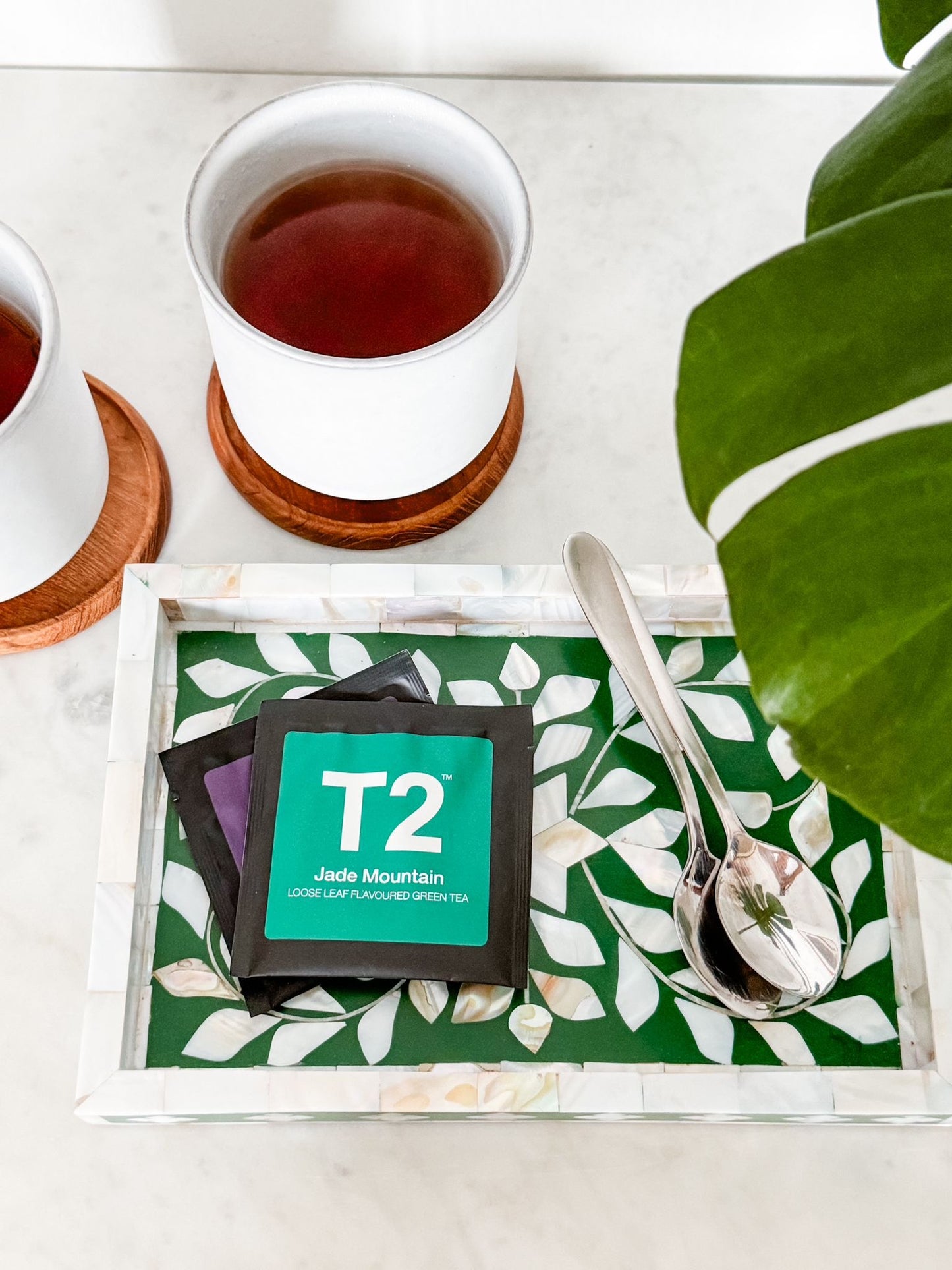 Tea leaves and T2 tea packaging on a green leaf-patterned coaster with a white cup of tea in the background.