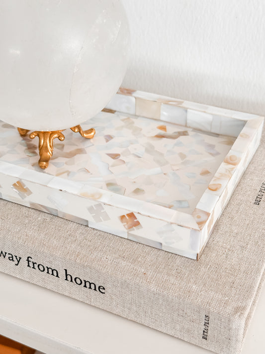 Decorative tray with shell pattern on a book titled 'Far from Home' with a white lamp in the background.