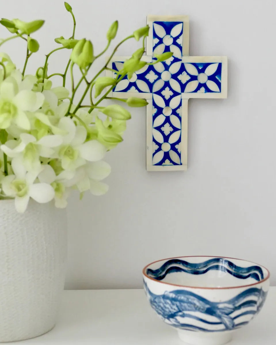 Decorative cross with blue and white pattern next to a vase with green flowers and a matching bowl on a white surface.