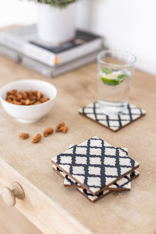 Set of four black and white patterned coasters on a wooden surface with a glass of water and bowl of nuts.