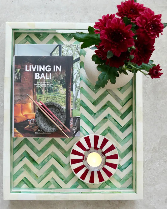 Decorative candle holder with chevron pattern, book titled 'Living in Bali', and red flowers on a gray surface.