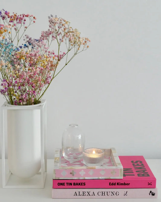 Vase with flowers, books, and a candle on a white surface in pink tray