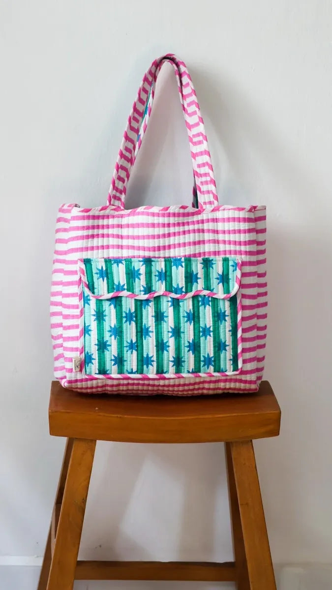 Tote bag with pink and white striped handles and green and white patterned front pocket on a wooden stool.