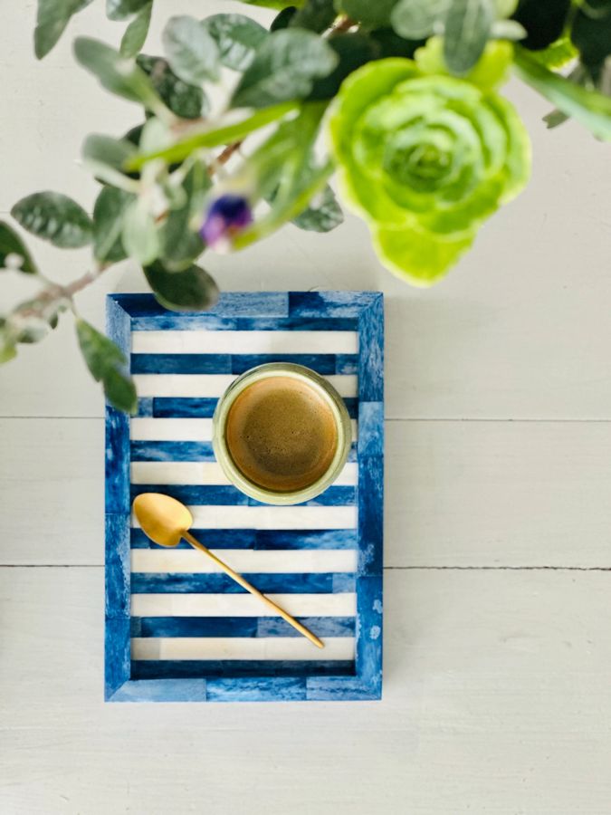 Blue and white striped tray with a gold cup and spoon on a light wooden surface with greenery in the background