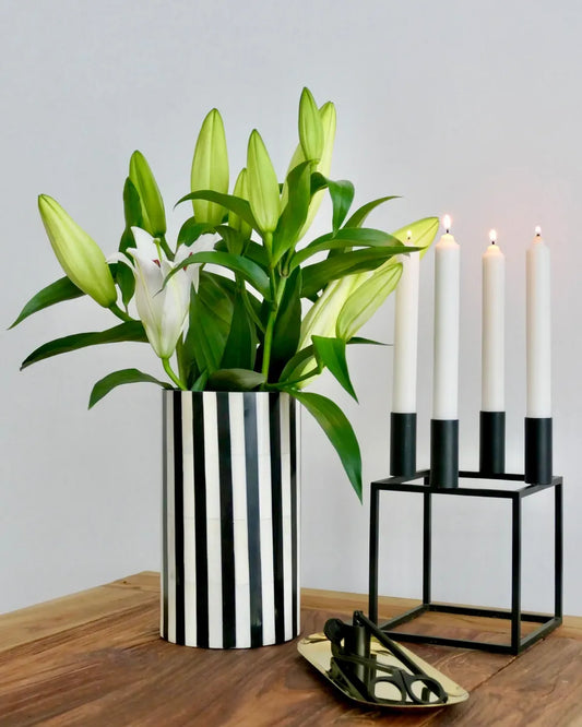 Striped vase with green plant and white flowers next to a black candle holder with three candles on a wooden surface.