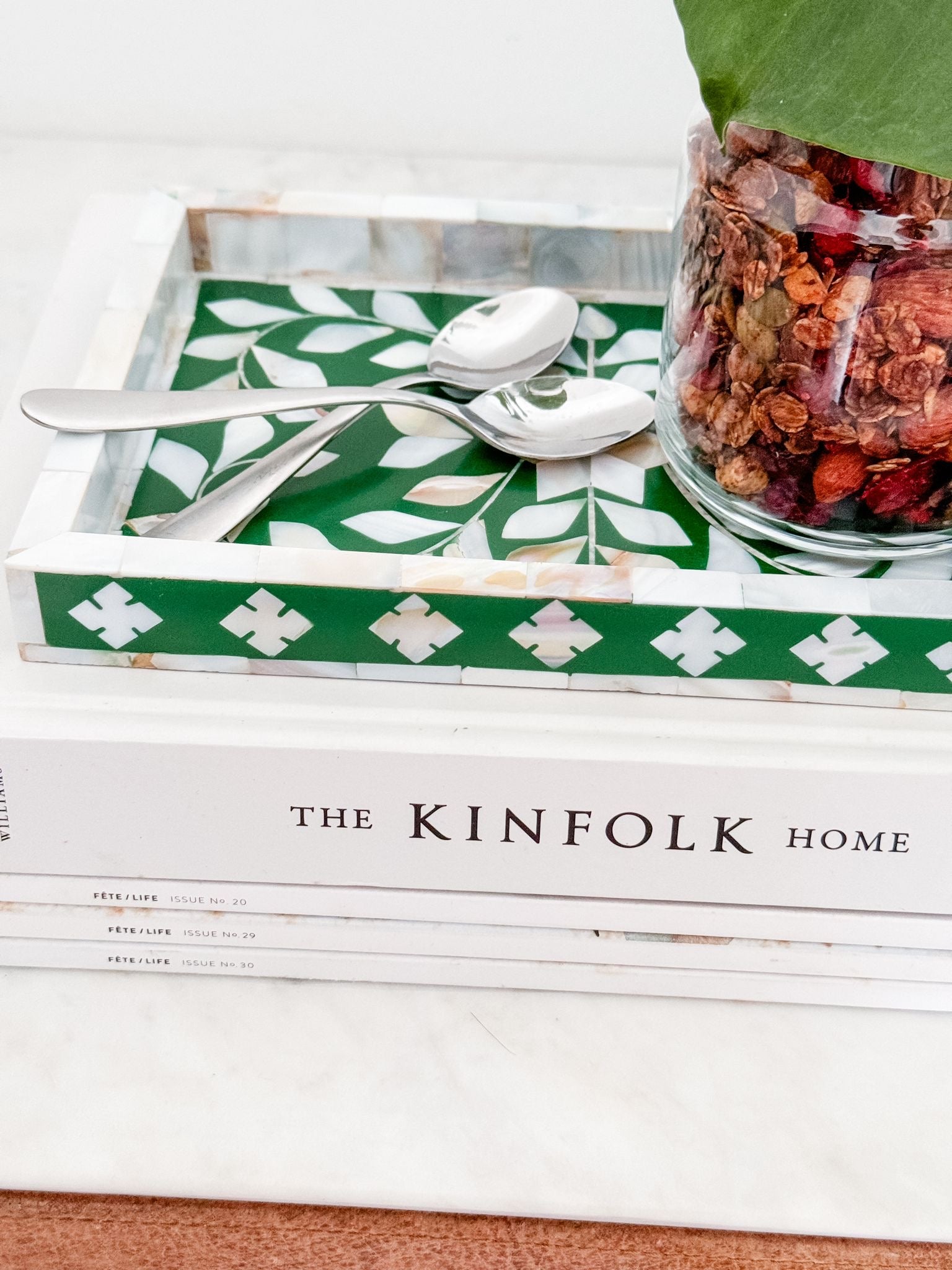 Stack of 'The Kinfolk Home' books with a green and white patterned tray, silver spoons, and a jar of dried fruits on a white surface.