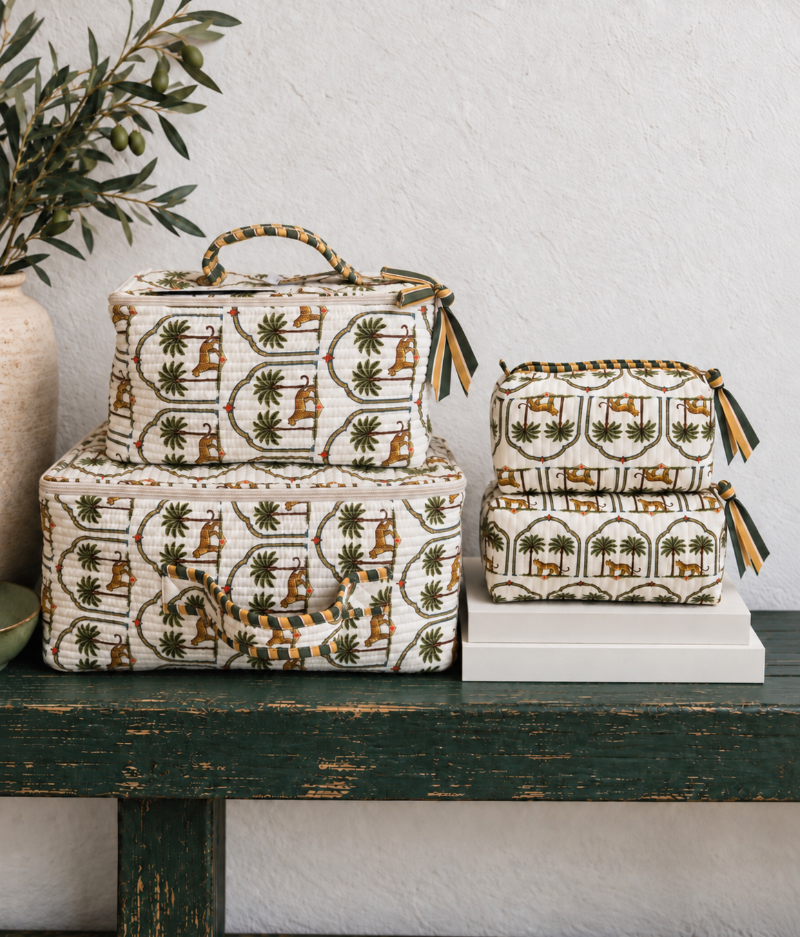 A set of fabric storage bags and suitcases with a vintage-style camel and palm tree print, styled on a rustic dark green wooden bench against a textured white wall.