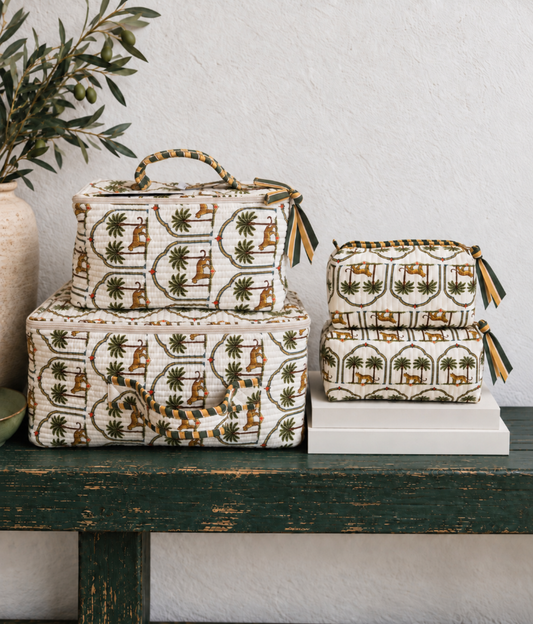A set of fabric storage bags and suitcases with a vintage-style camel and palm tree print, styled on a rustic dark green wooden bench against a textured white wall.