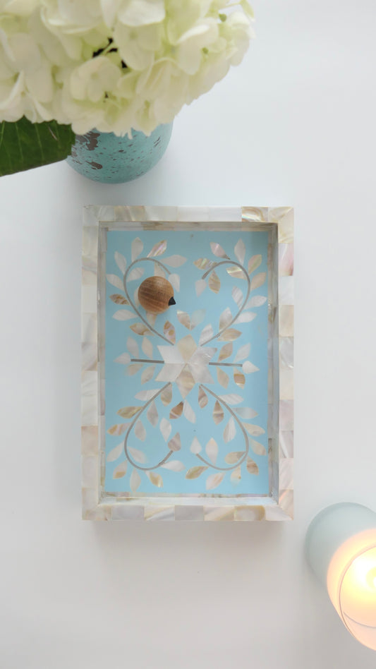 Decorative wall art with a shell and leaf design on a light blue background, placed on a white surface with flowers and a candle.