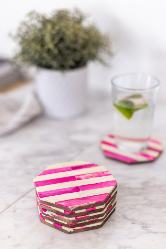 Hexagonal coasters with pink and brown stripes on a marble surface with a glass of water and plant in the background.