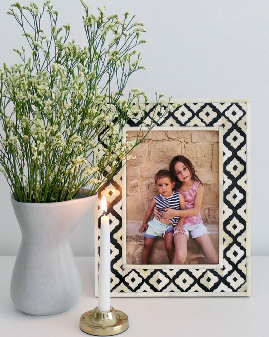 Decorative photo frame with a black and white pattern, holding a photograph of two children, next to a vase with flowers and a candle.