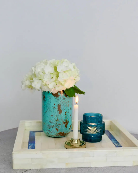 Decorative setting with a turquoise vase, white flowers, and candles on a tray against a light gray background.
