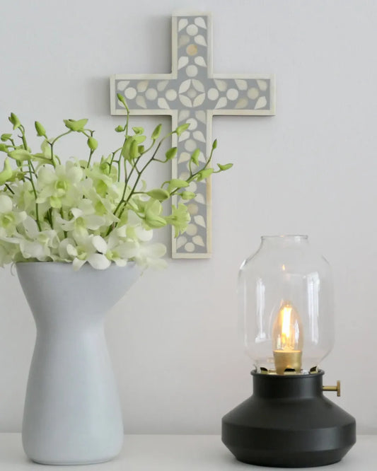 Decorative cross on a wall with a vase of flowers and a lamp on a table.