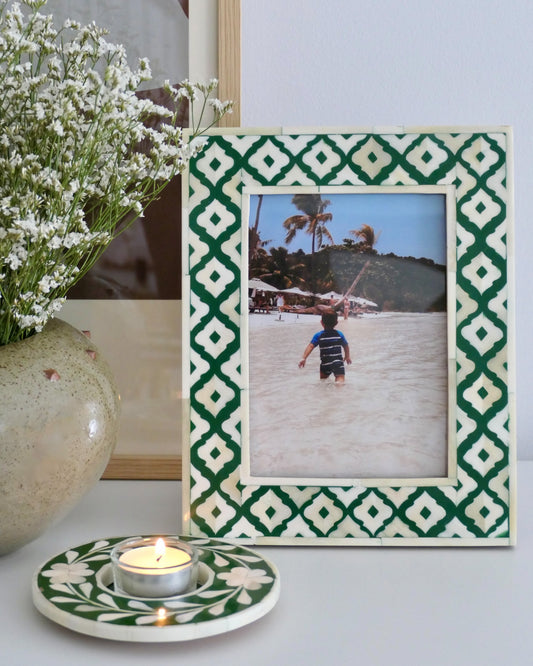 Decorative photo frame with green and white pattern, containing a beach photo, next to a vase with white flowers and a candle.
