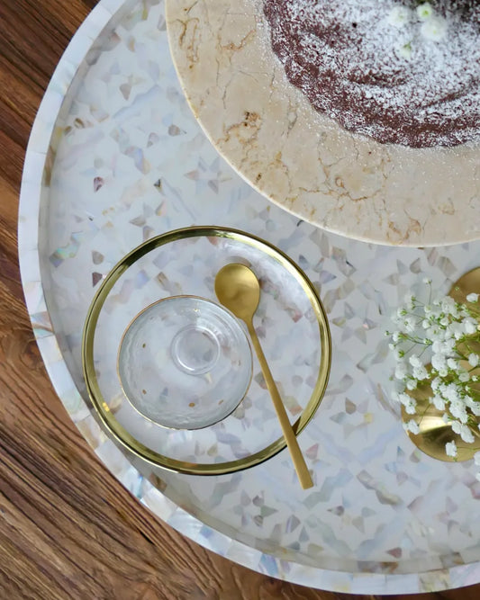 Glass dish with gold rim and spoon on a decorative tray with cake and flowers.